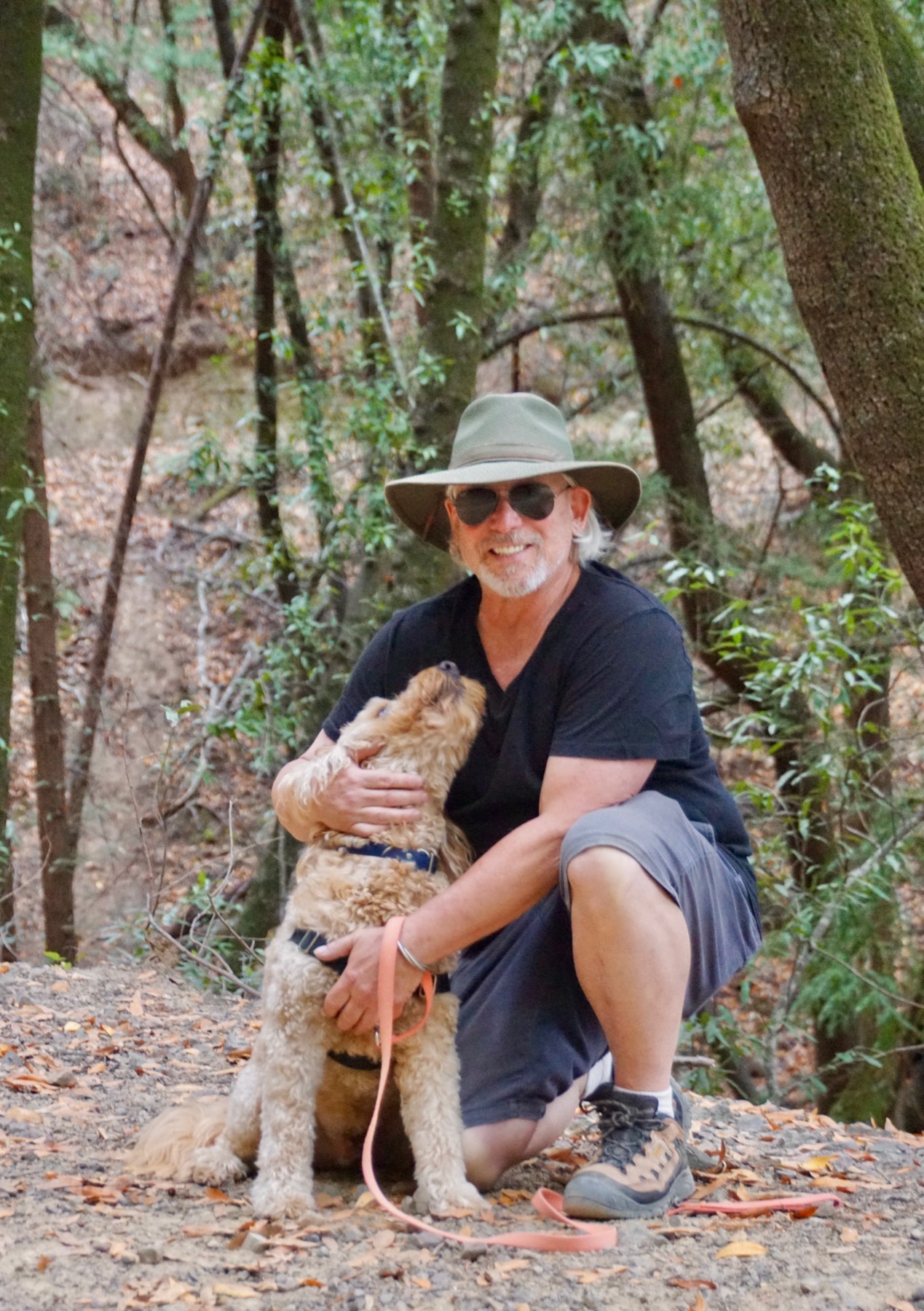 Gary Adler kneeling on a shaded fire road with his arm around a doodle looking up at him